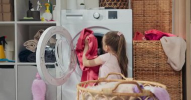 Küçük kız ev işlerine yardım etmeyi sever. Banyo zemininde oturuyor, kirli çamaşırlarını sepetinden çıkarıp çamaşır makinesine atıyor. Yanında çamaşır sepeti var..