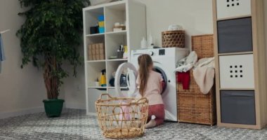Pembe elbiseli küçük güzel kız çamaşır makinesinden çamaşırları çıkarıp çamaşır sepetine koyuyor. Kız ev işi yapmayı seviyor. Giysileri katladı, sepeti aldı ve odaya taşıdı..