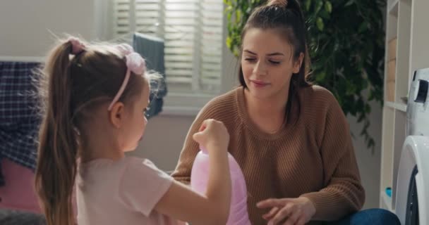 年轻的母亲和女儿在洗衣服 女孩手里拿着粉红洗衣粉 递给妈妈 女人拧开软木塞 和女儿一起闻到香气 他们在笑 在聊天 — 图库视频影像