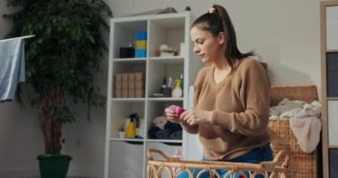 Genç anne, kızlarının elbiselerini yıkadı ve sepetten çıkardı. Çorap ve pembe elbise katlıyor. Kadın elbise tutuyor ve gülümsüyor. Arkada çamaşır makinesi ve banyo aksesuarlarıyla dolu bir raf var..