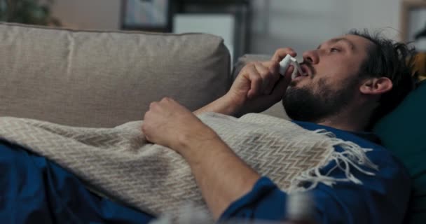 Человек Бородой Лежит Диване Покрыт Одеялом Брюнетка Прижимает Спрей Горлу — стоковое видео