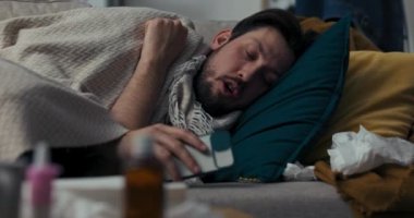 Genç adam battaniyeyle kaplı olarak kanepede yatıyor. Esmer çok öksürüyor, kendini iyi hissetmiyor. Adam telefonu elinde tutuyor, gözlerini kapıyor ve uykuya dalıyor. Yanında peçete ve haplar var..