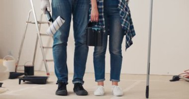 Ayakta duran, elinde boya silindiri ve kova tutan kadın ve erkeğin bacakları. Kot pantolon, siyah beyaz spor ayakkabı giyiyorlar. Kadının yüzüğü var. Arkasında merdiven ve duvar boyama malzemeleri var..