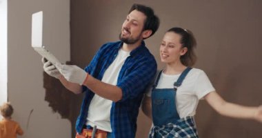 Karı ve kocanın aileleriyle görüntülü görüşmeleri var. Evde tamirat yapıyorlar, duvarları boyuyorlar ve ne yaptıklarını gösteriyorlar. Mavi gömlekli sakallı adam dizüstü bilgisayar tutuyor. Arkasında, oğlum duvarları boyuyor..