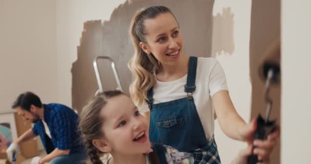 家庭正在整修房屋和粉刷墙壁 拥有卷发的母亲向女儿展示如何正确地画墙壁 他们紧紧地抱在一起 谈着话 微笑着 爸爸在后面画墙 — 图库视频影像