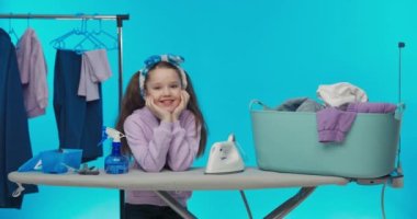 Renkli mavi bir stüdyo arka planında çamaşır odası. Küçük gülümseyen tatlı kız ev işlerini yapıyor, çamaşırları sıralıyor, ütüye hazırlanıyor..