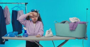 Küçük bir kız stüdyoda mavi bir arka plana karşı duruyor. Çocuk burun tıkama pis kokulu giysiler ütü.