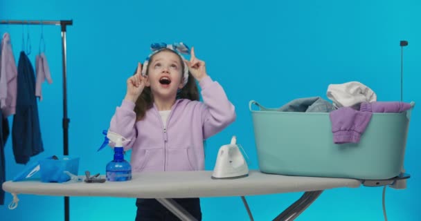 讽刺的衣服 快乐的微笑着的女孩指着一个空白的广告向上指着 喜欢做家务活 拿着熨斗 折叠着一件衬衫 — 图库视频影像