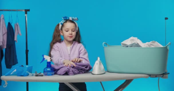 头戴头巾的性感小女孩和两只站在蓝色粉刷工作室背景上的小马从大碗里拿出衣服 准备熨烫衣服 — 图库视频影像
