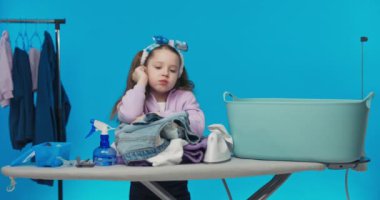 Aşırı çalışan bir kız kameraya üzülerek bakar, ütü masasına eğilir, giysileri ütüler, çamaşırları hazırlar, mavi bir duvara yaslanır. Günlük ev işleri..