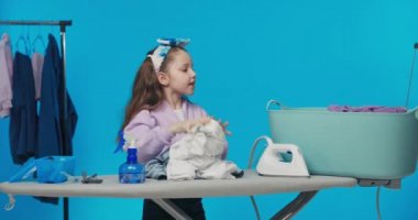 Saç bandı takmış küçük tatlı bir kız ve iki midilli mavi pastel stüdyonun üstünde duruyor. Arka planda giysileri katlıyor, büyük kaseden çıkarıp ütülemeye hazırlanıyor..