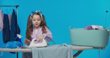 Güzel küçük kız ütülüyor giysiler, elindeki işe odaklanıyor gülümsüyor, demiri dikkatlice kullanıyor, ev işleri, mavi arka plan..
