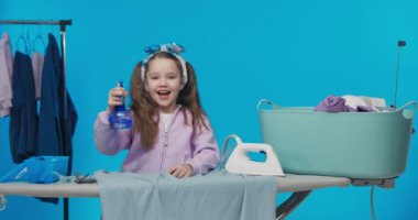 Küçük temizlikçi kadın ev işlerine yardım eden çalışkan bir kızdır. Sevimli çocuk elinde ütü makinesi ve demir su kabıyla mavi stüdyo arka planında duruyor..