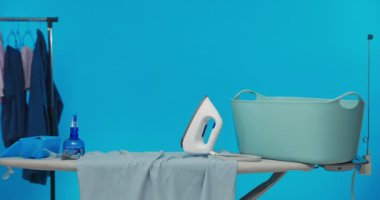 Küçük tatlı bir kız ütü masasının arkasından eğilip kıyafetlerinin arkasına saklanıyor. Mavi stüdyo arka planı.