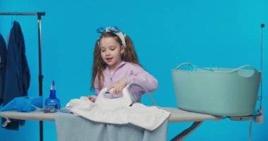 Güzel küçük kız ütülüyor giysiler, elindeki işe odaklanıyor gülümsüyor, demiri dikkatlice kullanıyor, ev işleri, mavi arka plan..