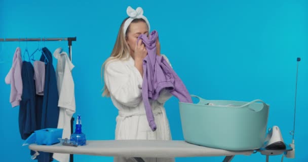掃除中にアイロンをかける準備をしていると 新鮮な下着のきれいな匂いの服を嗅ぐ女性 スタジオでの青の背景に笑みを浮かべて女の子の肖像 — ストック動画