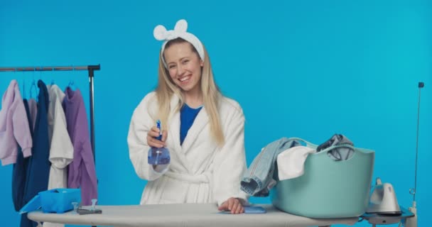 一位穿着浴衣的金发美女正在早上洗衣服 熨烫衣服 手里拿着一瓶喷雾水 工作室背景为蓝色的家庭主妇画像 — 图库视频影像