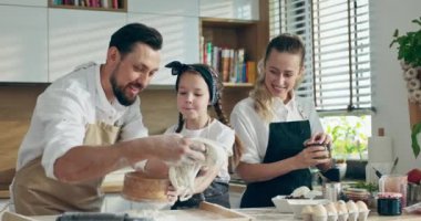 Genç anne pizza hazırlama sürecini izlerken, meraklı anaokulu öğrencisi kız modern hafif mutfakta babasıyla birlikte un elemeye yardım ediyor.
