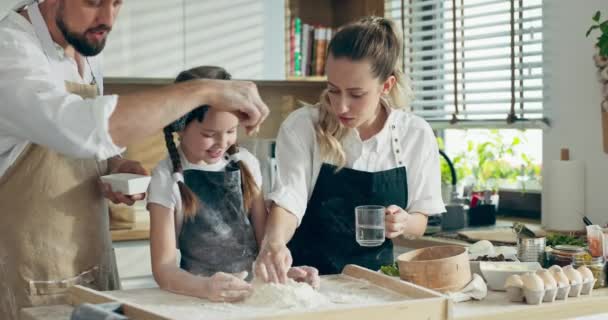 快乐的家庭观念 快乐的学龄前女儿在围裙帮助父母揉面 可敬的妻子把水倒在木制桌子上 而父亲则在挑食 烹调概念 — 图库视频影像