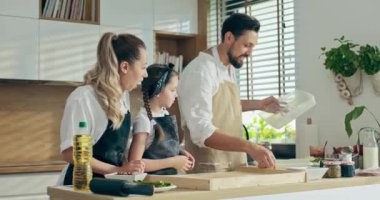 Pişirme işlemi. Genç, güzel bir anne, küçük bir anaokulu kızı ve modern mutfakta ev yapımı pizza için hamur yoğuran sevimli bir kocası var. Kızım ahşap yüzeye un dökmeye yardım ediyor.