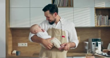 Modern mutfağın ahşap masasında duran sakallı genç baba yeni doğmuş oğlunu kucağında bebekle oynarken aile yemeği hazırlıyor.