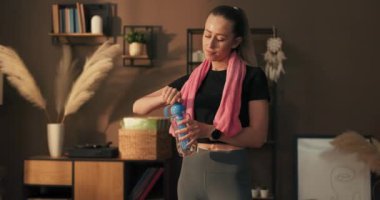 Güzel, güçlü, mutlu, genç bir kadın evde spor yapıyor. Boynunda havluyla bir kız odanın ortasında durup antrenmandan sonra dinleniyor..