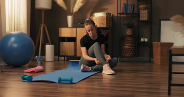一个女人在家里准备锻炼 坐在地板上的垫子上 穿上运动鞋 系上鞋带 舒适的鞋袜 — 图库视频影像