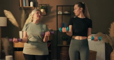 Mutlu genç kız hemşire kız kameraya bakıyor ve tatbikat yapıyor. Sevimli yaşlı kadınla ısınıyor. Kadınlar dambıllarla spor kıyafetleri giyerek antrenman yapıyor..