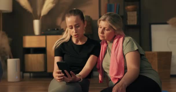 若いトレーナーと彼女の主人公はヨガマットの上に座って 練習のインスピレーションを実行するために携帯電話で見て トレーニングシリーズの間に休んで — ストック動画
