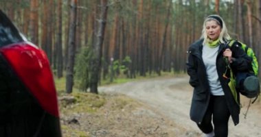 Ormanda yürüyen yaşlı bir kadının fotoğrafı. Arabasına geri geliyor. Uzun bir yolculuktan sonra paspaslı spor sırt çantasıyla ormanda yürüyüş yapıyor..