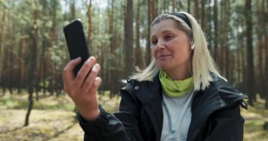 Sarışın büyükannenin kafasında bandajla ormanın ortasında elinde bir telefonla otururken çekilmiş yakın plan fotoğrafı. Yorucu bir yolculuktan sonra ailesiyle konuşurken elinde bir telefonla gülüyor. .