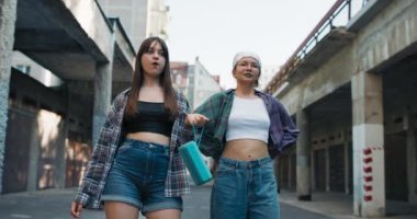 Güzel kıvırcık saçlı iki genç kız ellerinde müzik dinlemek için taşınabilir bir hoparlörle şehirde bir yerleşim bölgesinde yürüyorlar. Kızlar özgüvenle birbirlerine bakıyorlar ve ilerliyorlar..