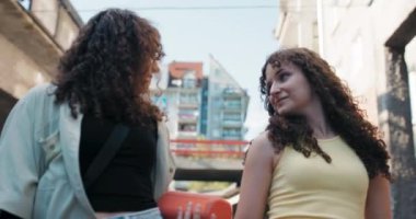 Güzel kıvırcık saçlı iki genç kız ellerinde müzik dinlemek için taşınabilir bir hoparlörle şehirde bir yerleşim bölgesinde yürüyorlar. Kızlar özgüvenle birbirlerine bakıyorlar ve ilerliyorlar..