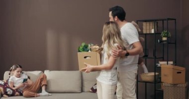 Anaokulu kızı rahat bir kanepede uzanmış cep telefonuyla oynarken anne ve babası yeni dairenin ortasında ellerinde saksıda saksılar ve ev eşyaları olan karton kutular tutuyorlardı. .