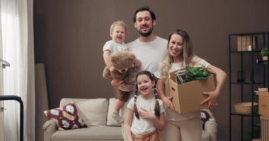 Sevinçli anaokulu kızı ve oğlu modern bir dairede kameraya bakıp gülerek saksı ve ev eşyalarıyla dolu kartpostallar taşıyorlar..