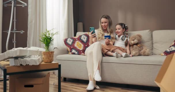 在现代宽敞宽敞的新公寓里 快乐的年轻妈妈和学龄前的孩子坐在舒适的沙发上 年轻女子手持智能手机 手握信用卡 键入电话号码 — 图库视频影像