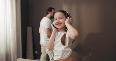 Mutlu meraklı anaokulu kızı kulaklık takıp rahat koltukta müzik dinlerken ebeveynler taşınan yeni daireleri dekore ediyorlar. Anne ve baba raflarda asılı..