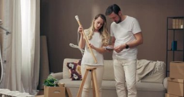 Genç, tapılası bir anne ve baba okuma talimatları ile arka planda saksı bitkileri ve ev eşyalarıyla döşenmiş yeni lamba döşenmiş rahat aparat kutuları ile dekore etmeye çalışıyor..