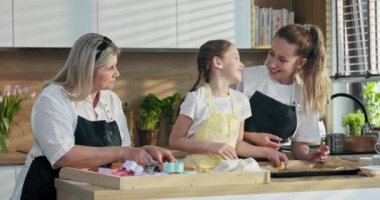 Orta yaşlı mutlu sarışın anne ve kızları modern mutfakta ev yapımı bisküvi pişiriyorlar. Anaokulu kızı yemek hazırlarken ablasını ve annesini öpüyor..