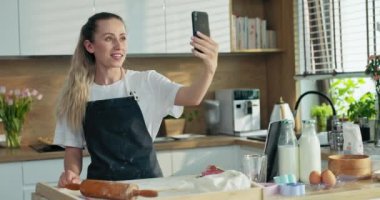 Sevinçle gülümseyen genç anne yuvarlanan ev yapımı hamurla merdane kurabiyeleri hazırlıyor pizza makarnası modern mutfakta konuşuyor kız ve oğluyla video bağlantısı ile konuşuyor.
