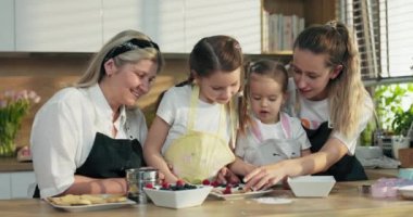 Anneler Günün kutlu olsun. Mutlu orta yaşlı bir büyükanne, kızı ve sevimli anaokulu torunları ev yapımı kurabiyeleri böğürtlen ile giydirip modern aydınlık mutfakta önlükle duruyorlar..