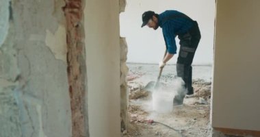 Takım elbiseli, beyzbol şapkalı bir adam tadilat altındaki bir daireyi temizliyor. Duvarları kürekle yıktıktan sonra zeminden arta kalan enkazı topluyor..