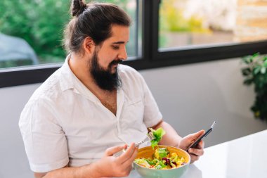 Mutlu genç adam evde çalışırken ve cep telefonuna bakarken yemek molası veriyor.
