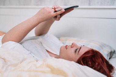 Genç bir kadın gülümsüyor, cep telefonuyla selfie çekiyor, yatak odasında uzanıyor..