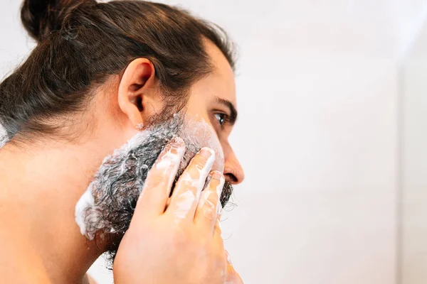 Gros plan d'un jeune homme avec une barbe touffue, mettant de la crème à raser sur sa barbe. — Photo