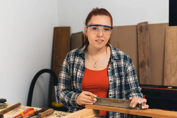 Genç kızıl saçlı bayan marangoz, küçük marangozluk atölyesinde hassas bir seviye kullanıyor. Kameraya bakıyorum.