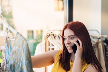 Genç kızıl saçlı kadın bir moda dükkanından kıyafet alıyor. Kadın cep telefonuyla konuşuyor. alışveriş kavramı.