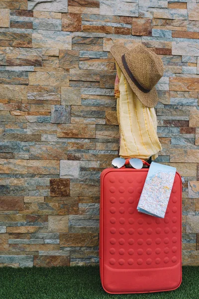 Luggage in front of a stone wall and grass floor to use text. suitcase with hat, sunglasses, scarf and map. — Foto de Stock