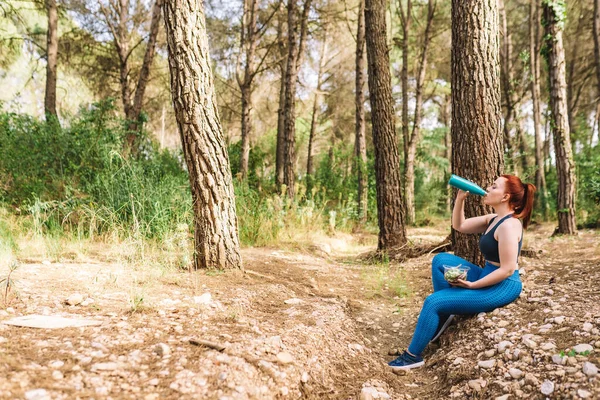 young sporty woman sitting in a forest resting after training. sporty girl doing training exercises. health and wellness lifestyle. copy space, outdoor public park, natural sunlight.
