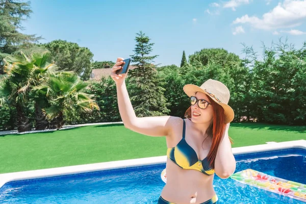 young girl in bikini, taking a selfie in the pool at home with her smartphone. young woman on summer holiday sunbathing at home. concept summer and free time. outdoor garden, natural sunlight.